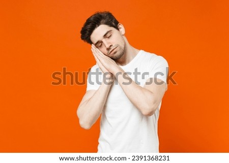 Relaxed handsome young man 20s wearing basic casual white blank empty t-shirt sleep with folded hands under cheek keeping eyes closed isolated on bright orange colour wall background, studio portrait