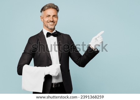 Adult barista male waiter butler man wear shirt black suit bow tie gloves elegant uniform point index finger aside on area work at cafe isolated on plain blue background. Restaurant employee concept Royalty-Free Stock Photo #2391368259