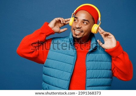 Young man of African American ethnicity wear padded vest red hat listen to music in headphones look aside on area isolated on plain dark royal navy blue background studio portrait. Lifestyle concept