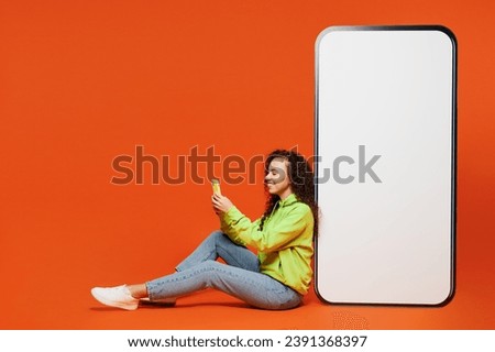 Full body sideways young woman of African American ethnicity in green hoody casual clothes sit big huge blank screen mobile cell phone with area use smartphone isolated on plain red orange background