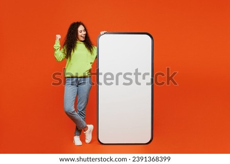 Full body young woman of African American ethnicity wear green hoody casual clothes big huge blank screen mobile cell phone smartphone with area do winner gesture isolated on plain orange background