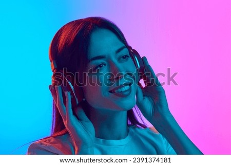 Young woman inn her 30s listening to music in headphones against gradient pink blue studio background in neon light. Enjoyment. Concept of human emotions, lifestyle, youth culture, facial expression Royalty-Free Stock Photo #2391374181