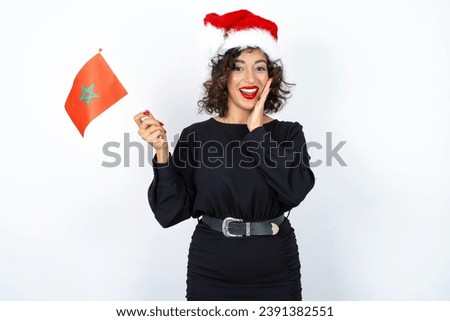 Young beautiful woman with curly hair wearing black dress, christmas hat and holding a Moroccan flag smiling and holding hand near face. 