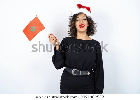 Young beautiful woman with curly hair wearing black dress and christmas hat over white studio background holding a Moroccan Flag Royalty-Free Stock Photo #2391382559