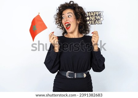 Young beautiful woman with curly hair wearing a black dress holding a Moroccan flag and a happy new year banner over white studio background.