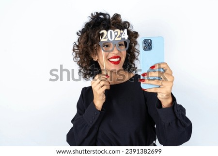 Young beautiful woman with curly hair wearing black dress, holding happy new year 2024 glasses and making a video call to friends and family with her smartphone .