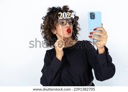 Young beautiful woman with curly hair wearing black dress, holding happy new year 2024 glasses and making a video call to friends and family with her smartphone blowing kiss.