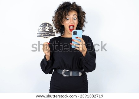 Young beautiful woman with curly hair wearing black dress, holding happy new year 2024 banner, holding phone and feeling surprised after received money transfer