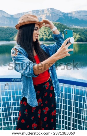 Smiling young female traveler in stylish bright clothing and hat taking selfie on mobile phone while standing near mountain lake during summer holidays.