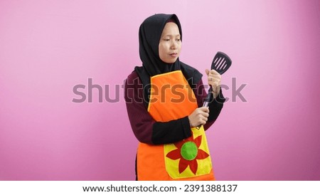 Beautiful smiling Asian woman wearing apron and hijab isolated on pink background. Household concept