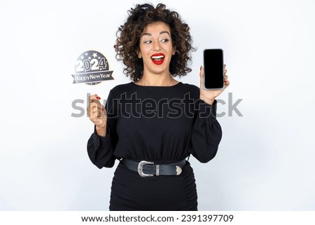 Young beautiful woman with curly hair wearing black dress, holding happy new year 2024 message showing blank screen picture. 