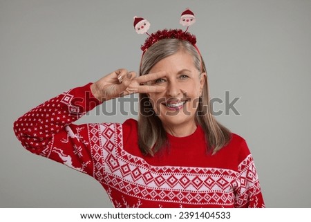 Happy senior woman in Christmas sweater and Santa headband on grey background