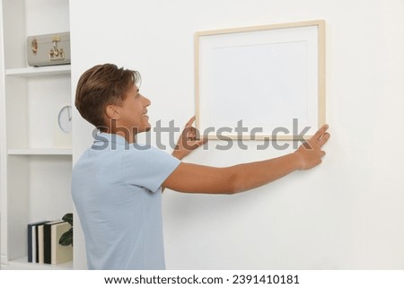 Young man hanging picture frame on white wall indoors