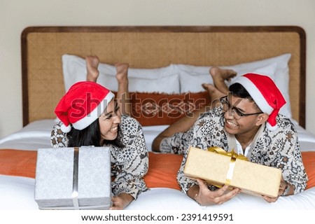 Happy young asian couple exchanging Christmas presents lying down on bed, wearing Santa hats celebrating holidays at home.