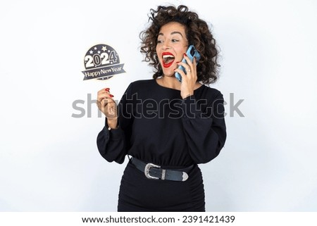 Young beautiful woman with curly hair wearing black dress, holding happy new year 2024 message and taking a picture or calling her family and friends via video call. 
