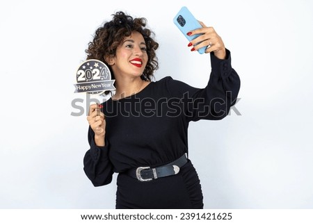 Young beautiful woman with curly hair wearing black dress, holding happy new year 2024 message and taking a picture or calling her family and friends via video call. 