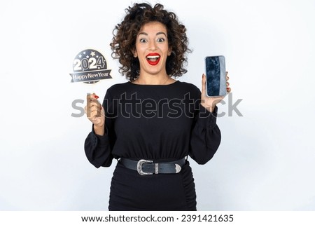 Young beautiful woman with curly hair wearing black dress, holding happy new year 2024 message showing blank screen picture. 