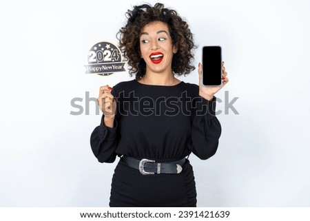 Young beautiful woman with curly hair wearing black dress, holding happy new year 2024 message showing blank screen picture. Woman looking aside at her phone.