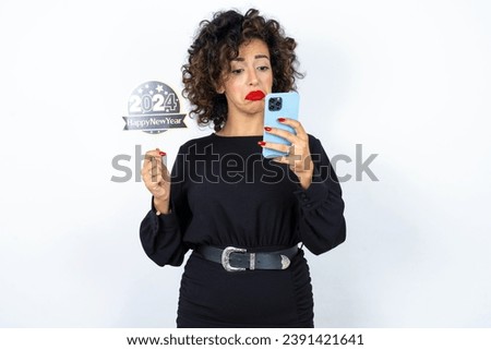Young beautiful woman with curly hair wearing black dress, holding happy new year 2024 message and taking a picture or calling her family and friends via video call feeling sad.  Royalty-Free Stock Photo #2391421641