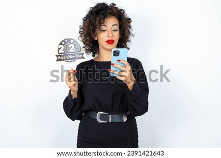 Young beautiful woman with curly hair wearing black dress, holding happy new year 2024 message and taking a picture or calling her family and friends via video call.