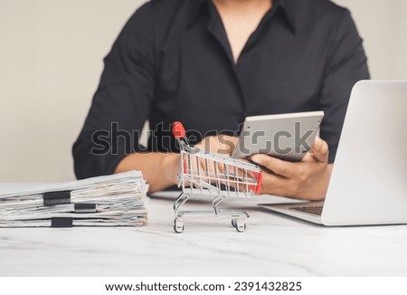 A small shopping trolley and a pile of documents on a table with a background of a woman using a calculator while sitting at the table in the office. Business and e-commerce concept. Close-up photo.