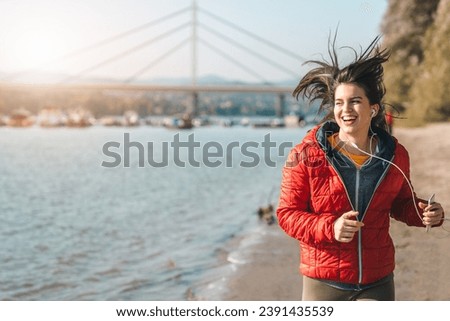 Young woman with toothy wide smile and wind in her hair running by the seaside surrounded by magnificent natural landscape.