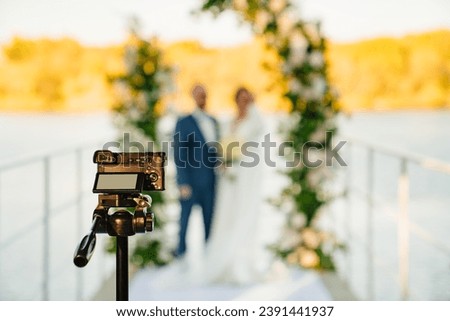 A video camera is filming the bride and groom at the off-site ceremony. Selective focus. Professional videographer and wedding photographer. Royalty-Free Stock Photo #2391441937