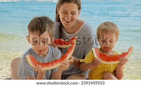 Mother and her two boys enjoy sweet watermelon on a beachside picnic. Family bonding during a holiday, summertime merriment, and the simple pleasures of a vacation.