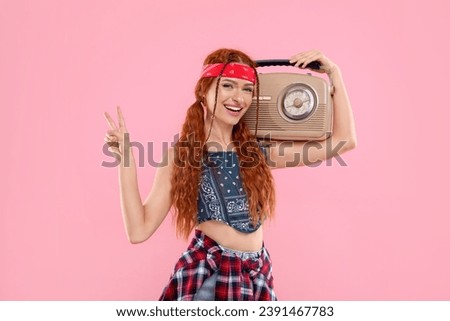 Stylish young hippie woman with retro radio receiver showing V-sign on pink background