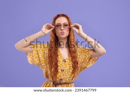 Stylish young hippie woman in sunglasses sending air kiss on violet background
