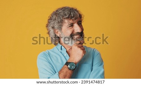 Close-up of an elderly gray-haired bearded man wearing a blue shirt, looking camera, thinking pleasant, smiling, isolated on an orange background in the studio Royalty-Free Stock Photo #2391474817