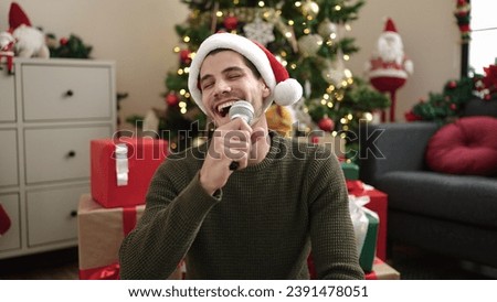Young hispanic man singing song sitting on floor by christmas tree at home Royalty-Free Stock Photo #2391478051