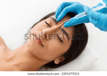 Beautician hands making hyaluronic acid injection for glabella facial rejuvenation procedure, young indian woman getting cosmetic injection between eyebrows at beauty center, top view Royalty-Free Stock Photo #2391479211