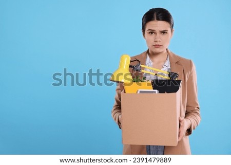 Upset unemployed woman with box of personal office belongings on light blue background. Space for text