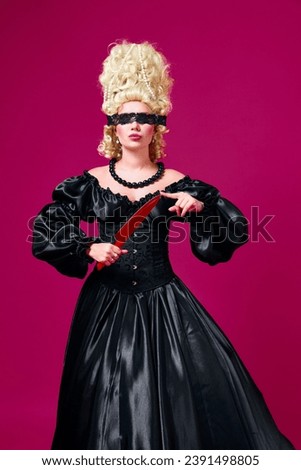 Portrait of medieval person dressed in old-fashioned black dress with openwork blindfold playing with color knife against magenta background. Concept of comparison of eras, shopping, style, fashion. Royalty-Free Stock Photo #2391498805