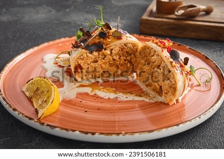 A tantalizing view of a cross-section of a stuffed squid, topped with vibrant garnishes and served beside a charred lemon wedge on a peach ceramic plate, against a dark grey background.