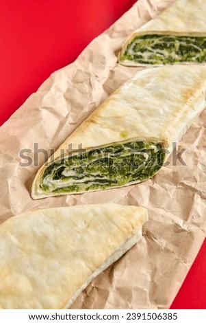 An angled view showcases the layers of a flaky spinach roll on brown parchment, with a bold red background accentuating the dish.