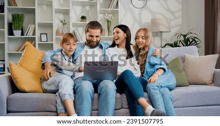 Happy family sitting together on couch in living room having fun on internet using laptop watch cartoons, parents buy goods for kids online. Smiling children their mother and father spend time