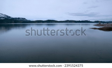 Long exposure shot of the lake at sunrise. Panorama view of the mountains and forest reflection in the water surface in an early morning with fog.  Royalty-Free Stock Photo #2391524375