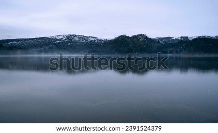 Long exposure shot of the lake at sunrise. Panorama view of the mountains and forest reflection in the water surface in an early morning with fog.  Royalty-Free Stock Photo #2391524379
