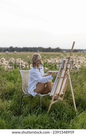 Girl artist, woman painting a landscape. Dressed in a white shirt, creating artistic creativity. Blank white canvas. Field in the background. Brush color palette paints. High quality shooting.