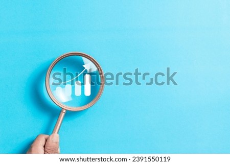 Hand holding magnifying glass focus to growth graph icon on blue background. Increase of chart positive indicators business. Analysis of investment strategies and economic trends.