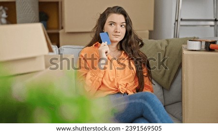 Young beautiful hispanic woman holding credit card sitting on sofa thinkin at new home