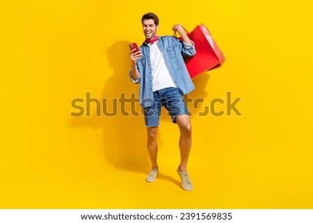 Full size photo of smart guy wear jeans jacket holding shopping bags read notification on smartphone isolated on yellow color background