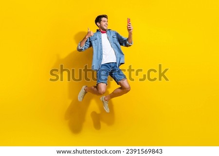 Full size photo of cool guy wear jeans jacket scarf jumping doing selfie show thumb up on smartphone isolated on yellow color background