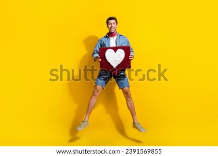 Full size photo of overjoyed guy wear jeans jacket shorts jumping hold heart tap like on comment isolated on yellow color background