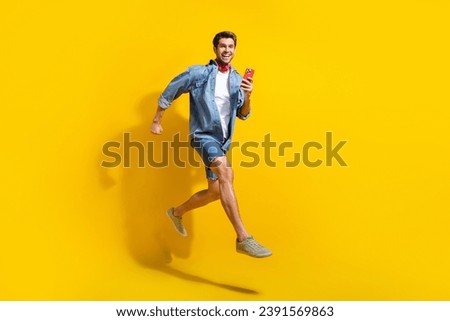 Full size photo of excited positive guy wear jeans jacket scarf run shopping holding smartphone in arm isolated on yellow color background