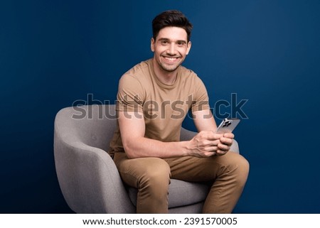 Photo of toothy beaming intelligent man dressed beige t-shirt sit in armchair with smartpone in arms isolated on blue color background
