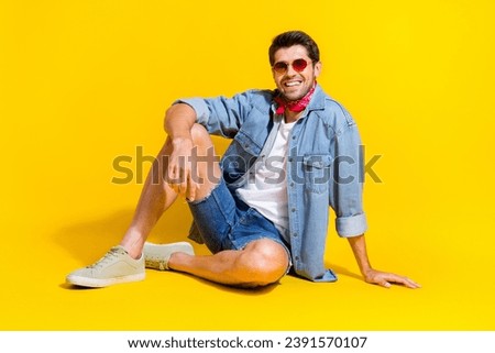Full size photo of positive cheerful stylish guy wear jeans jacket in sunglass sit on floor isolated on vibrant yellow color background
