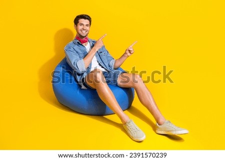 Full size photo of positive guy wear jeans jacket shorts indicating at logo empty space sit on pouf isolated on yellow color background Royalty-Free Stock Photo #2391570239